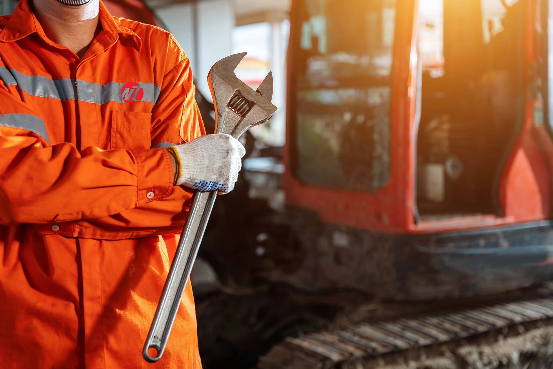 A person wearing orange work overalls and gloves holds a large adjustable wrench in front of a red construction vehicle with tracks, inside a building.