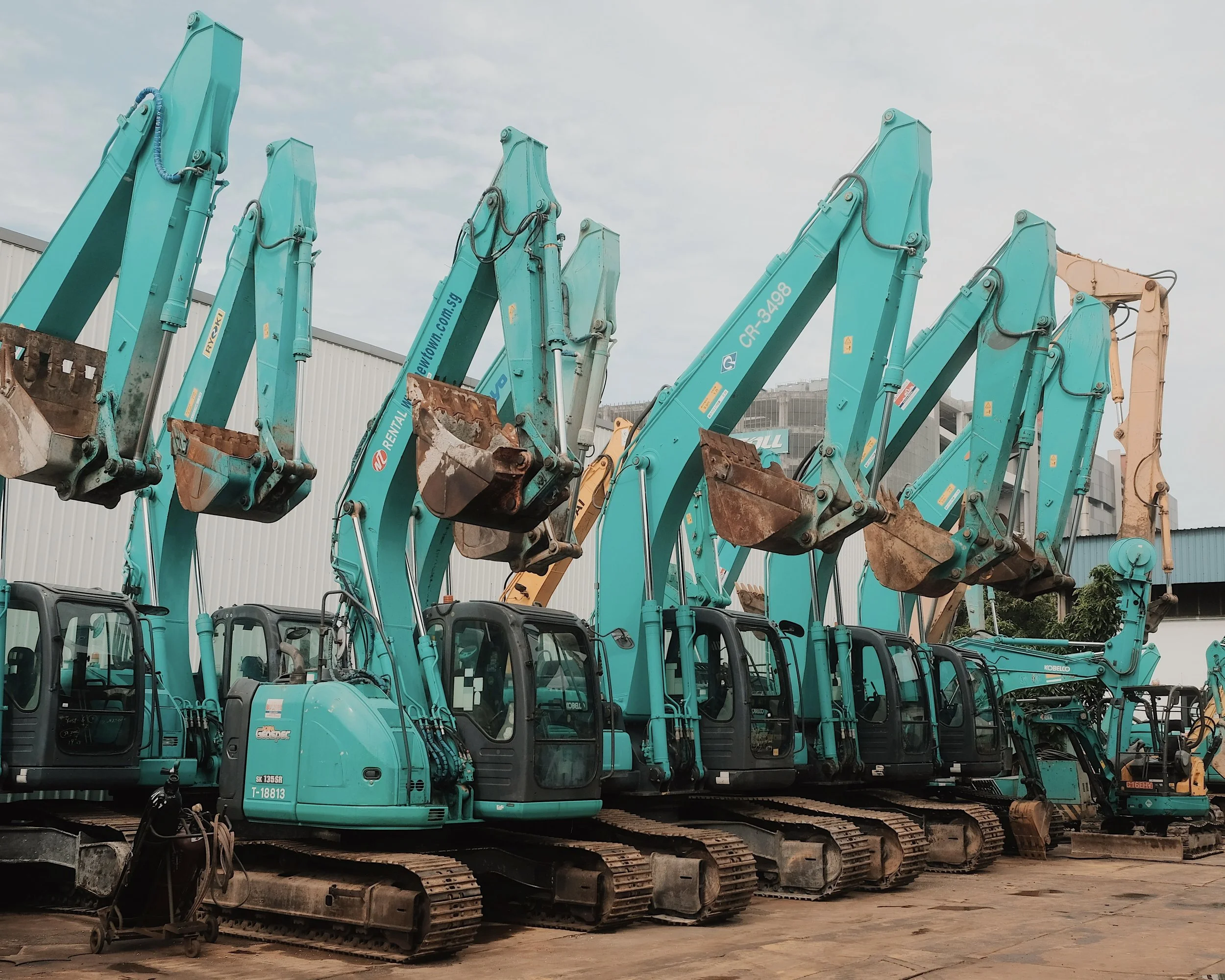 Multiple teal excavators lined up on a construction lot with a cloudy sky in the background.