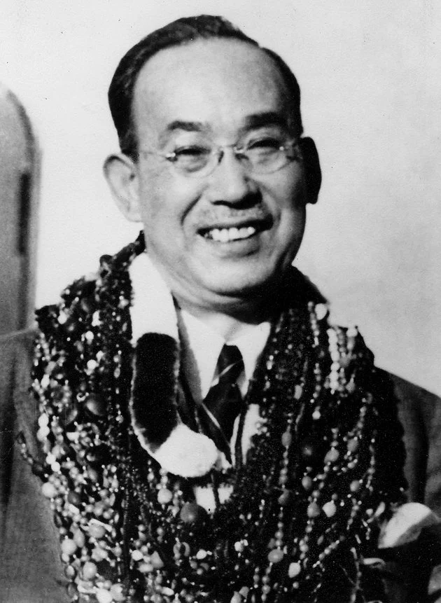 Black and white photo of a smiling man wearing glasses, a suit, striped tie, and multiple strands of necklaces around his neck.