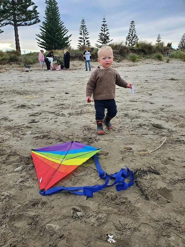 A young boy in a brown sweater and navy pants holding a string, standing on a sandy beach near a colorful rainbow kite on the ground. In the background, a group of people, some with children, are near tall pine trees and sand dunes under a cloudy sky