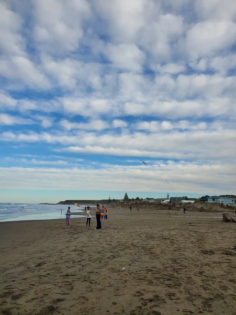 People flying kites on a sandy beach under a partly cloudy sky, with cliffs and buildings in the background.