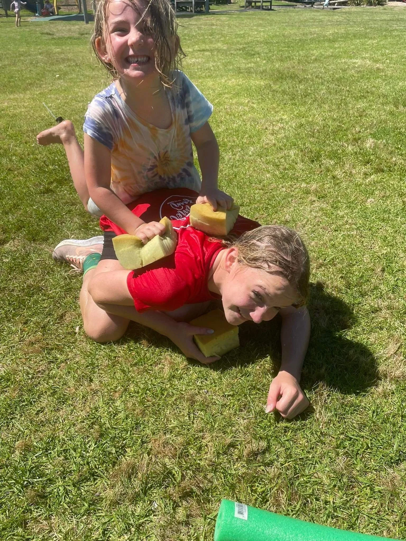 Two children playing on the grass, stacking sponges and lying on each other, outdoors on a sunny day.