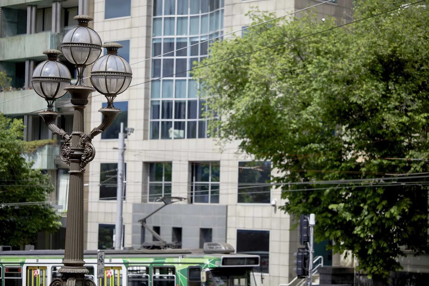 A vintage-style street lamp with four globed lights in an urban setting, with a modern building, a tree, and a tram in the background.