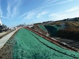 Construction site with a slope covered in green material, construction equipment, and a road in the background under a cloudy sky.