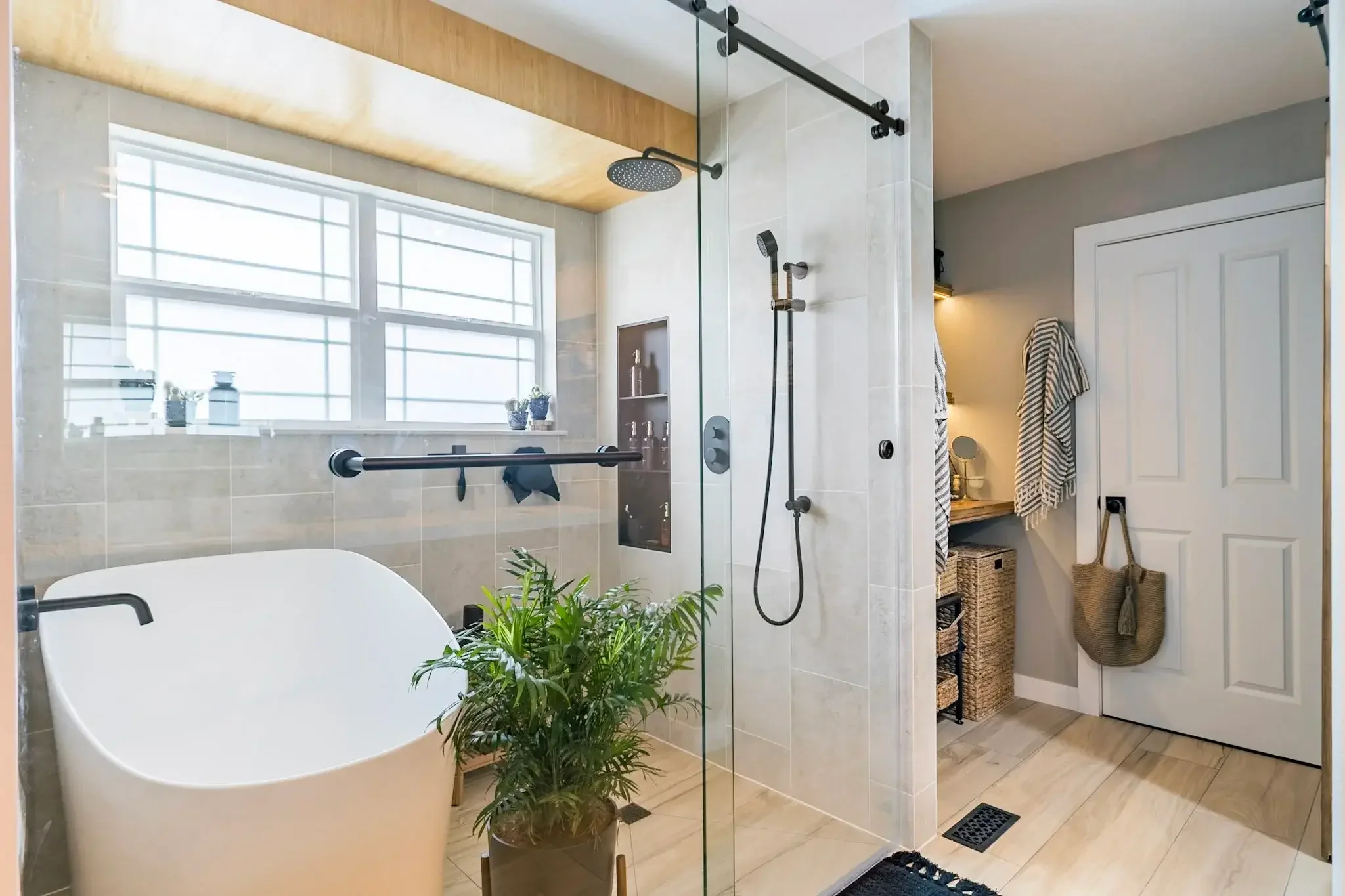 Modern bathroom with a freestanding bathtub near a large window, a walk-in shower with glass partition, and a small nook with towels and woven baskets. Green plants are placed for decor.