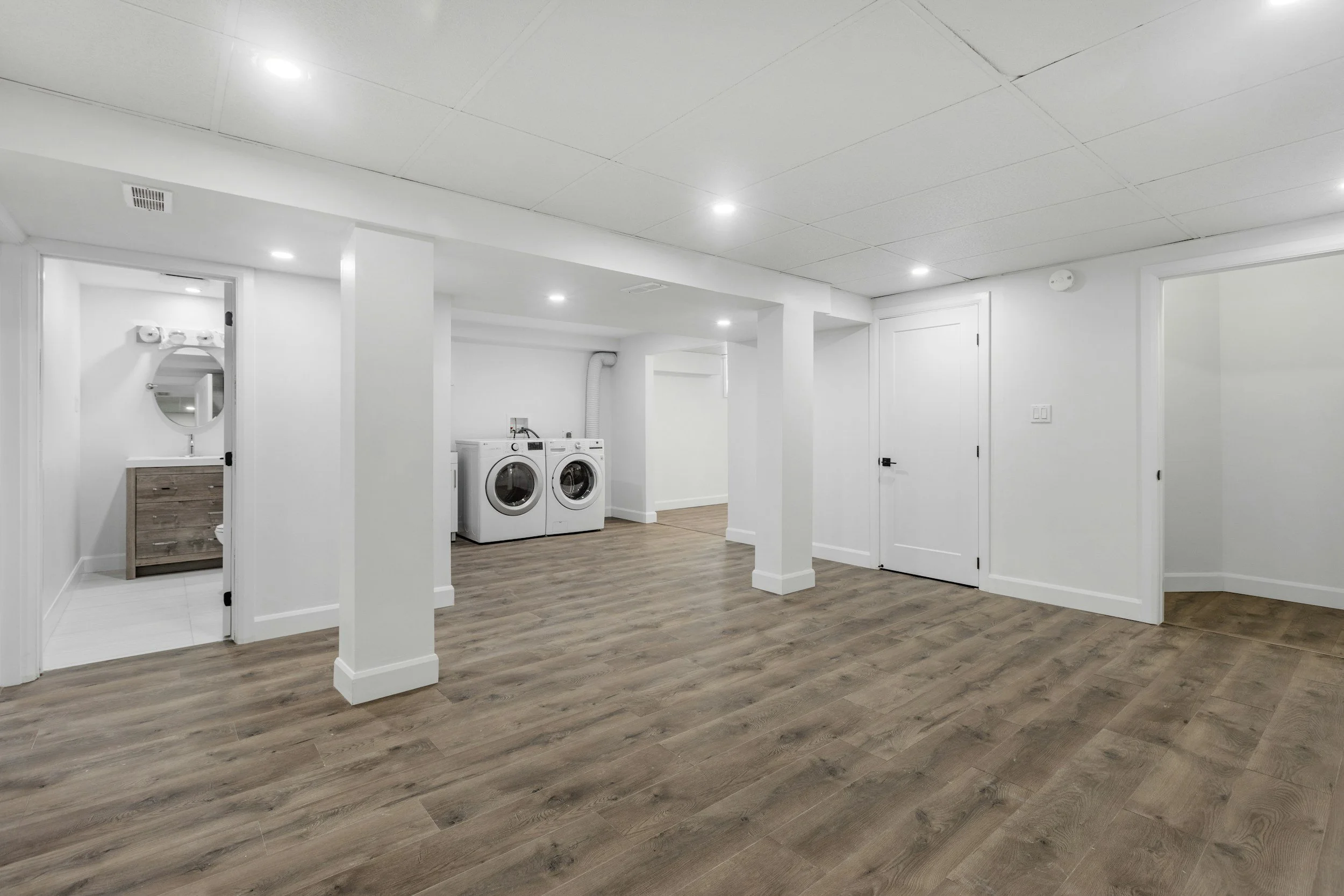 Empty finished basement with wood flooring, white walls, laundry area with washer and dryer, small bathroom with vanity, and multiple ceiling lights.
