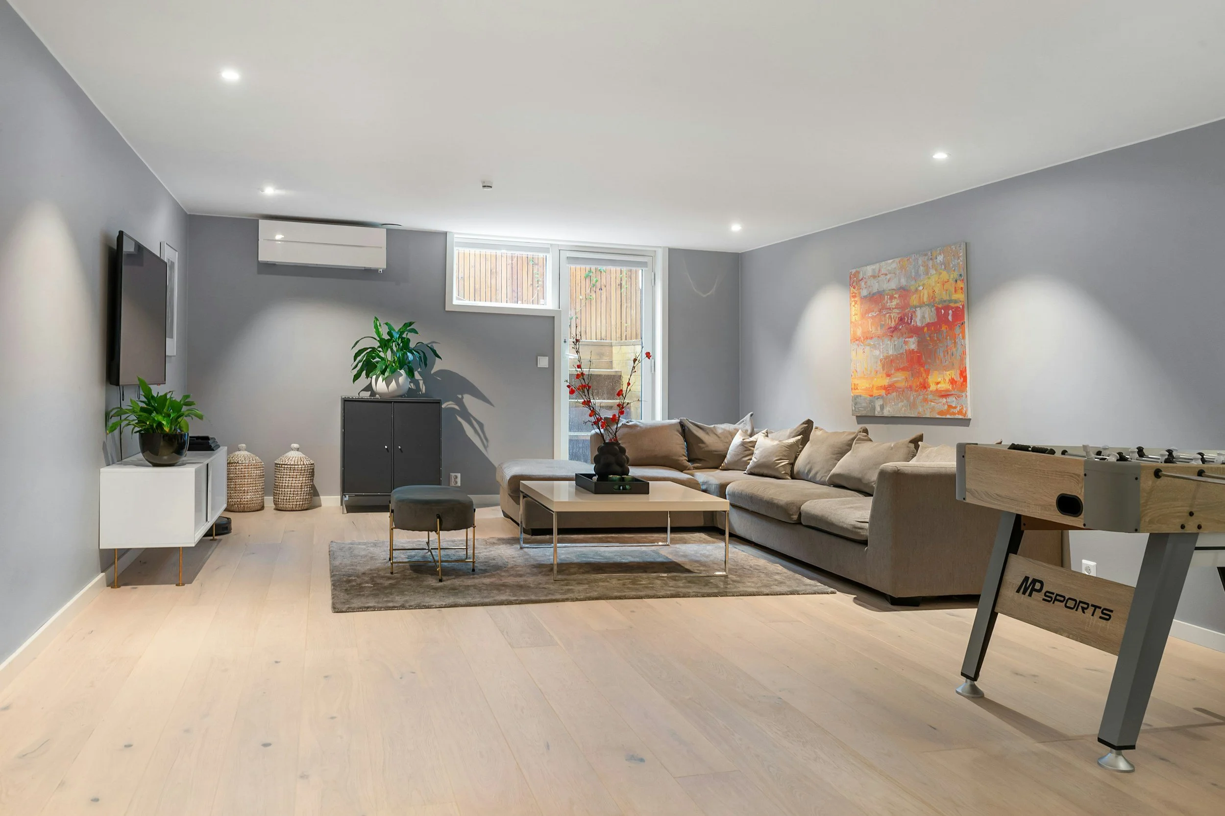 Living room with a beige sectional sofa, abstract orange and white painting, plants, small coffee table, small black cabinet, wall-mounted TV, foosball table, and an MP Sports workout machine, with light wood flooring and gray painted walls.