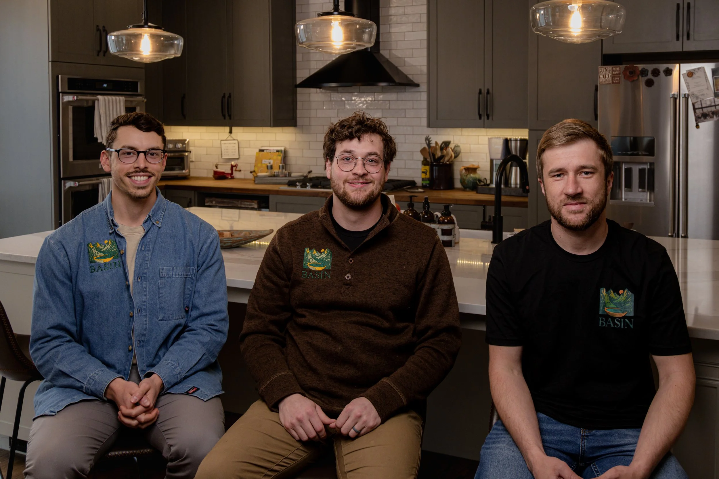 Three men sitting at a kitchen island, smiling at the camera. The man on the left wears glasses and a denim shirt, the middle man has curly hair and glasses, and the man on the right has short hair and a beard. All three wear dark shirts with a logo that says 'BASIN'.