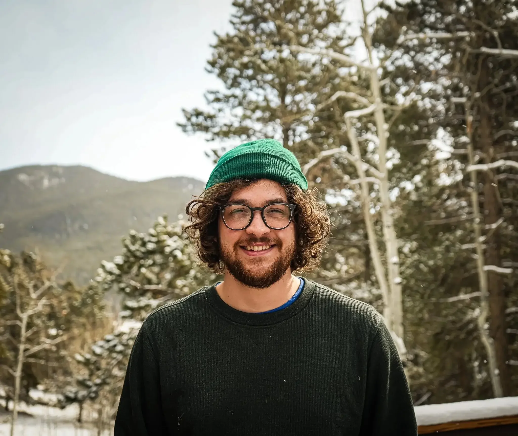 Close-up of a person with curly hair, wearing glasses, a green beanie, and a black sweater, outdoors with snowy trees in the background.