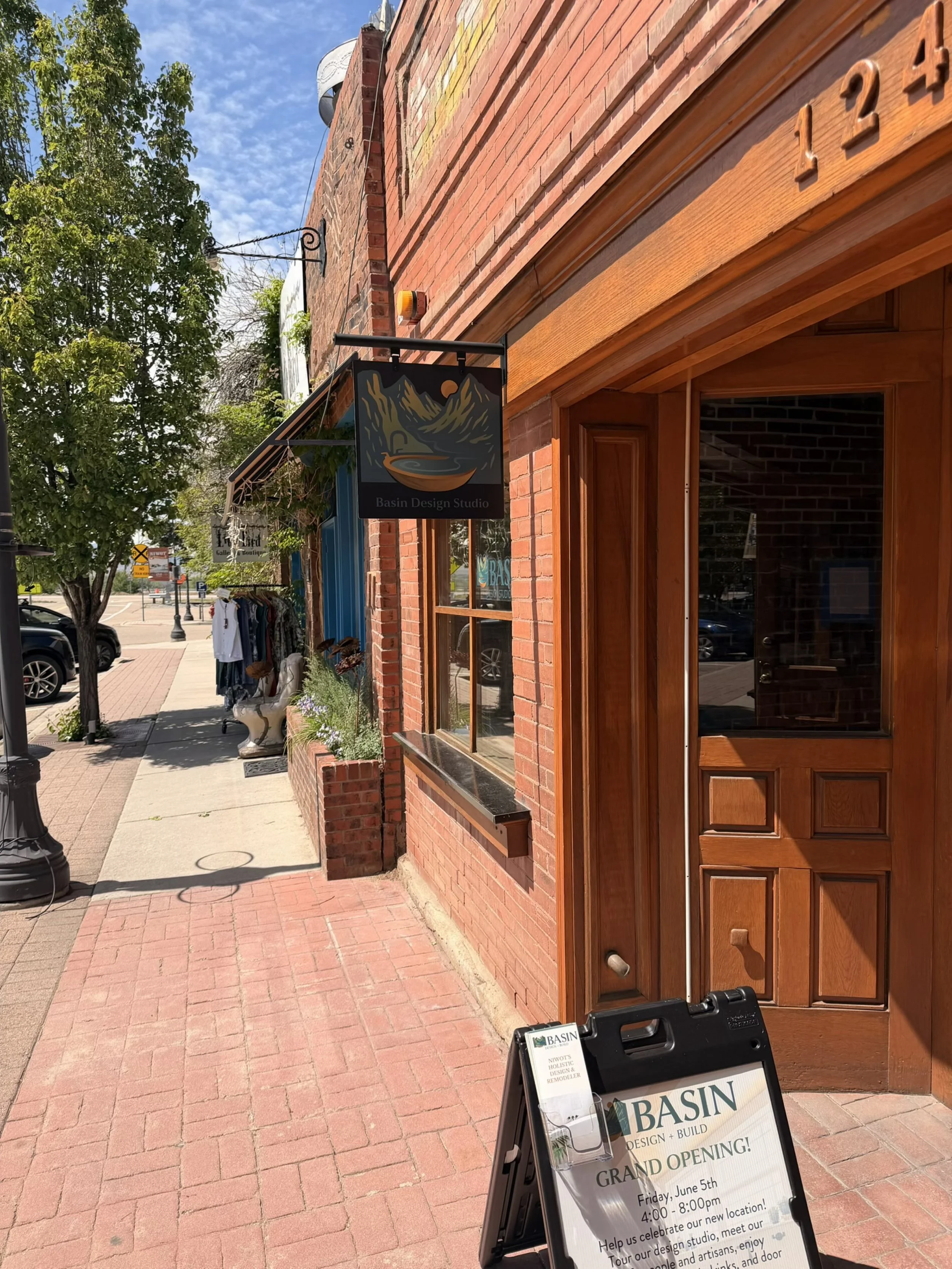 Street view of a brick storefront with a wooden door and windows. There are signs indicating a grand opening event for Basin Design Studio on June 5th from 4:00 to 8:00 pm, with information about studio tours and artisans. A chalkboard sign is placed on the sidewalk.