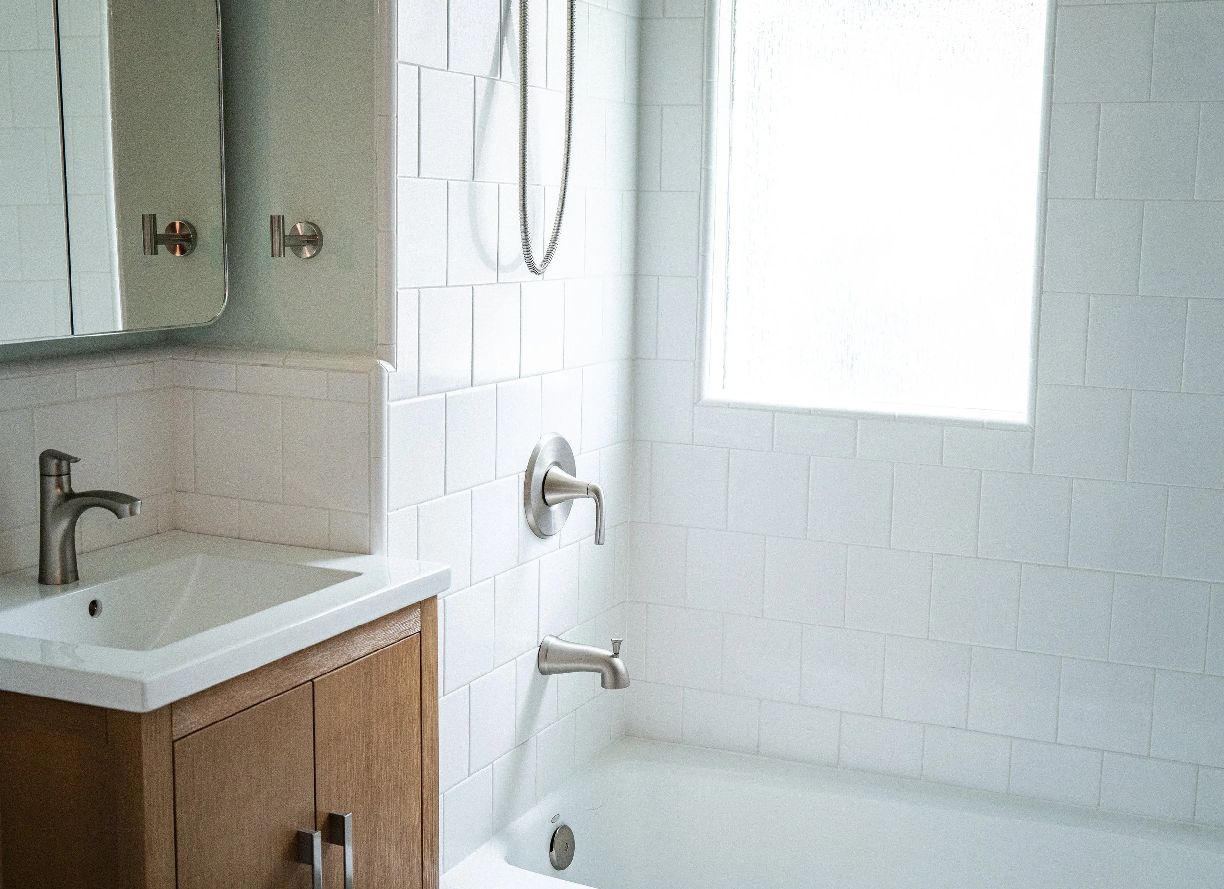 Modern bathroom with a white-tiled shower area, a small sink with a wooden cabinet, a mirror, and a large frosted window letting in natural light.