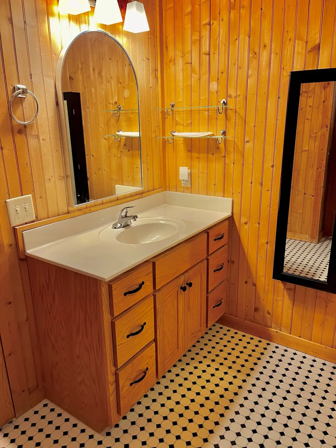 A bathroom with wood-paneled walls, a white countertop with a sink, a mirror, and a black and white patterned tile floor.