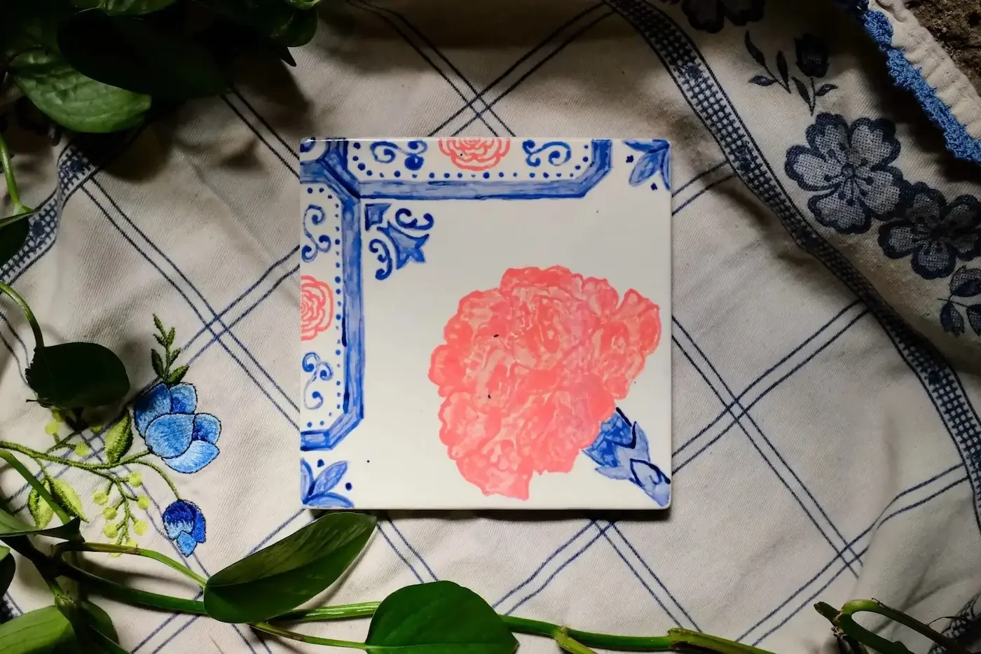 Custom hand-painted ceramic tile featuring a coral pink peony with a blue scrollwork border, styled on an embroidered floral linen with trailing pothos vines.
