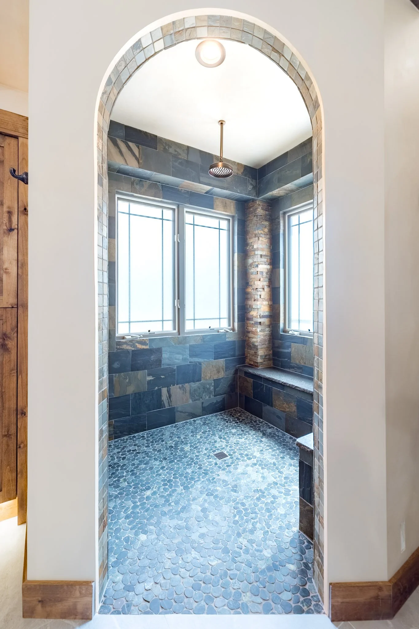 View of a walk-in shower with blue and gray slate tiles, pebble floor, brick column, and two windows, framed by a white arch with a tiled border.