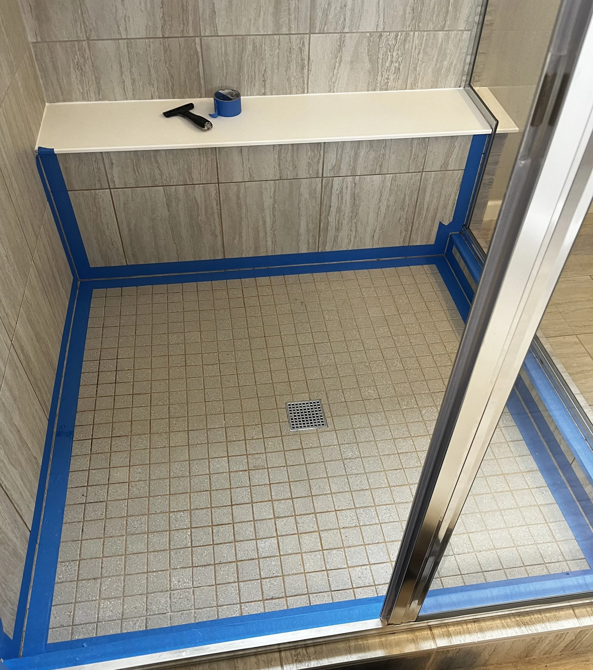 Empty bathroom shower stall with tiled walls and floor, blue tape around the edges, drain in the floor, and tools on a shelf.