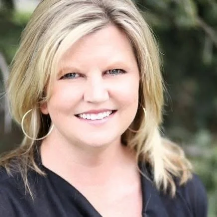 A woman with blonde hair, blue eyes, and a bright smile, outdoors with greenery in the background.