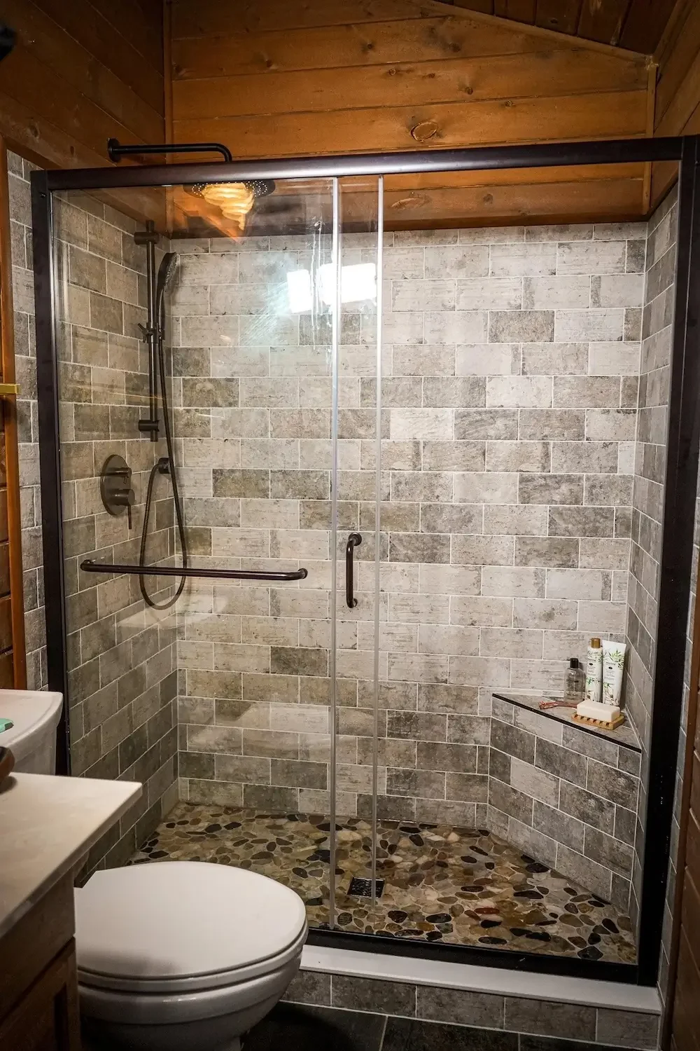 Bathroom remodel with walk-in glass shower, gray tile walls, built-in bench, and black fixtures.
