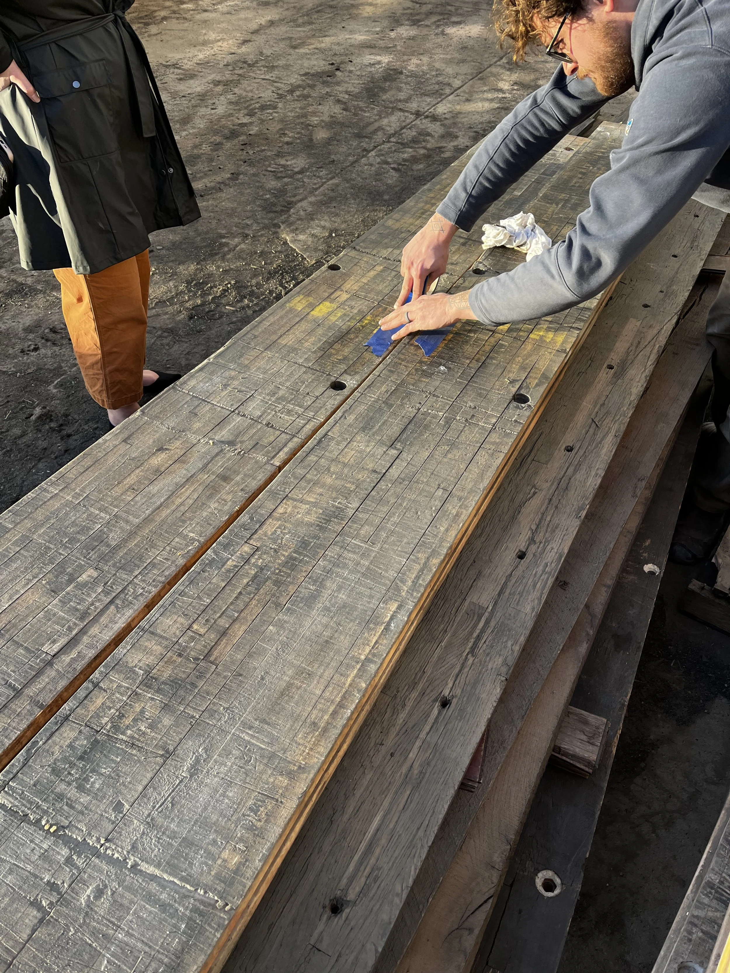 A man with curly hair, glasses, and a beard is working on a wooden workbench, using tape to mark or align a piece of wood, with a crumpled white cloth nearby. Another person stands nearby, partly visible in the image, wearing a black jacket and orange pants.