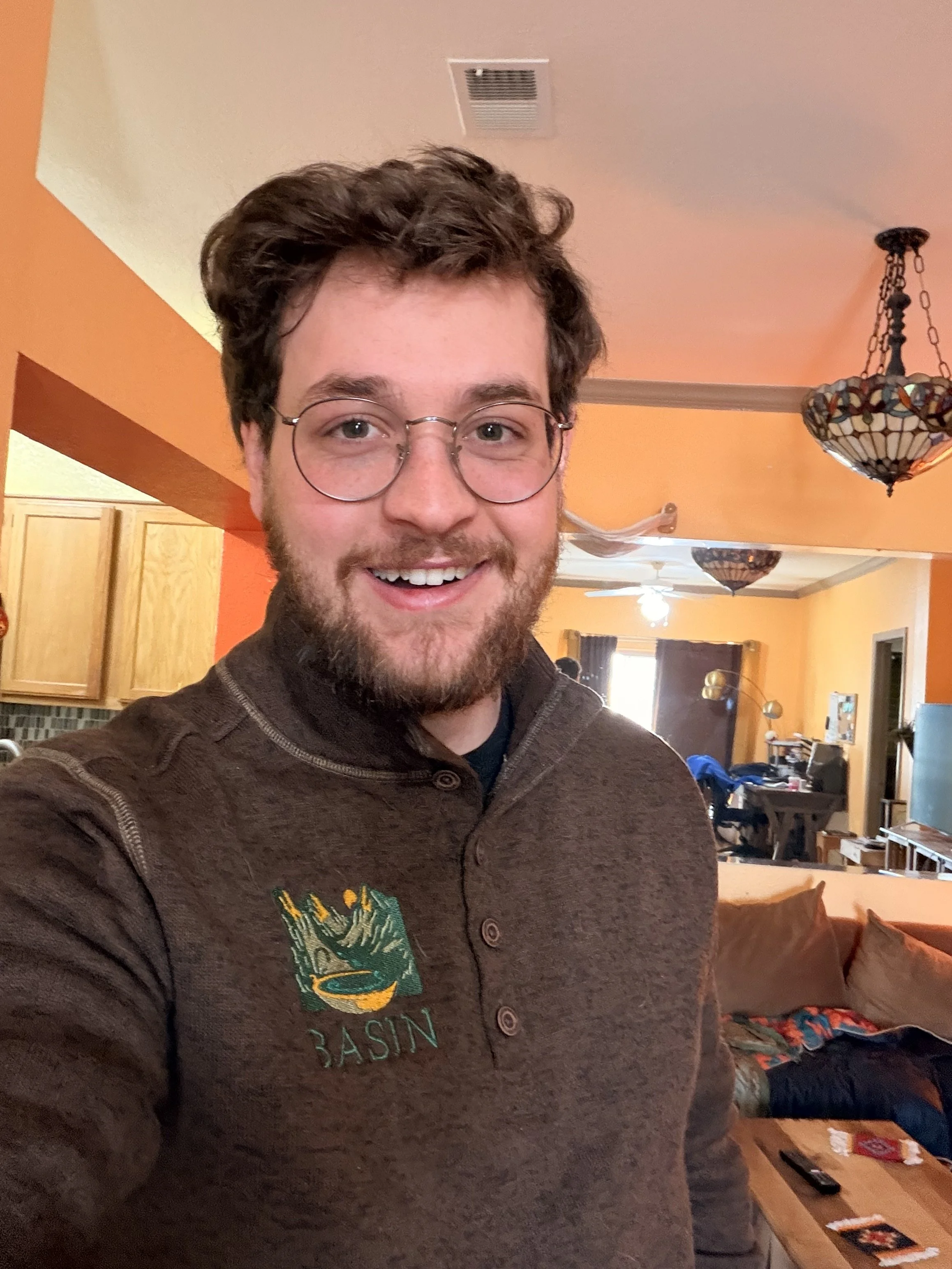 A smiling man with glasses and a beard taking a selfie in a cozy living room with orange walls, wooden cabinets, and pendant lights.