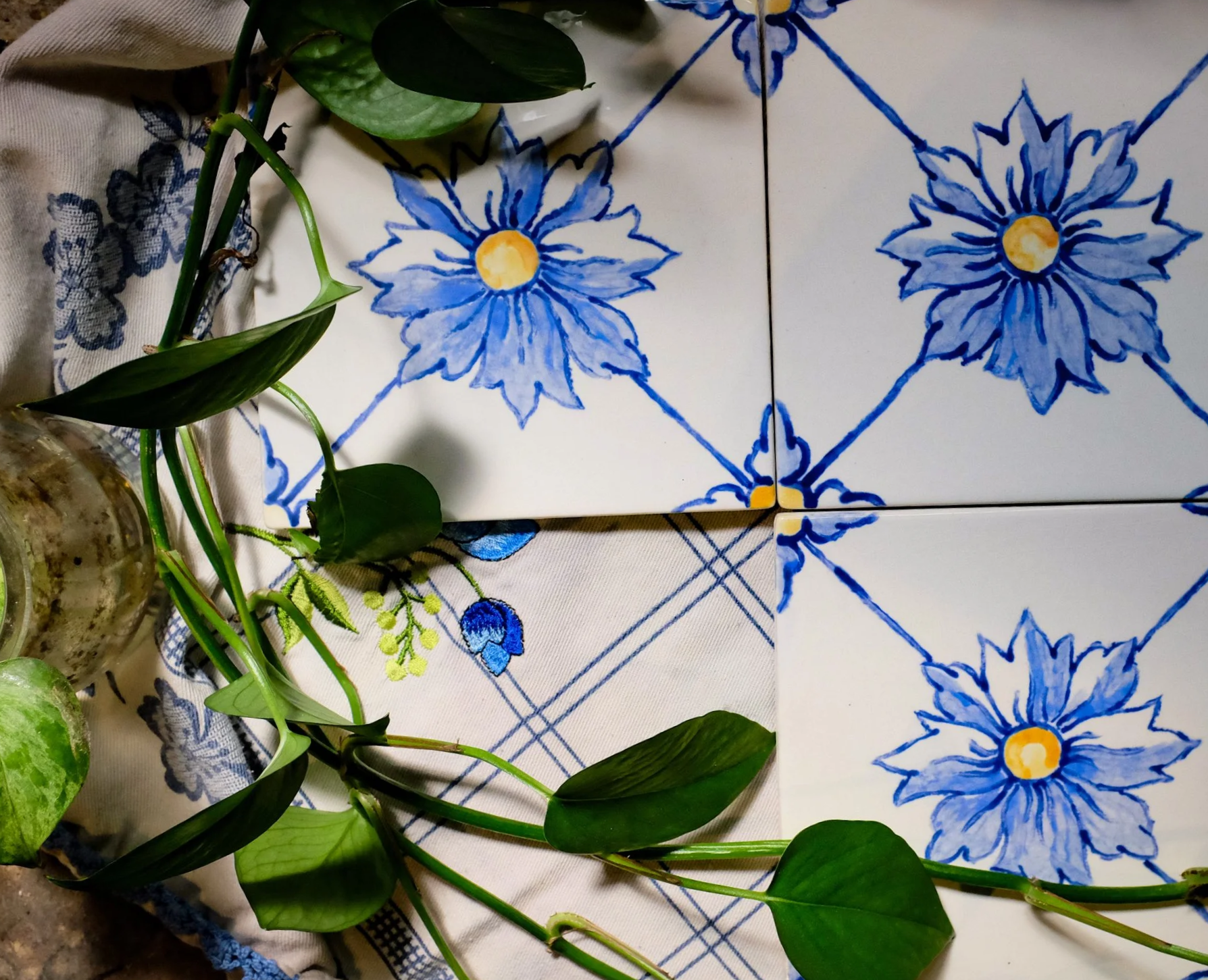 A photo showing a decorative tiled surface with a blue floral pattern, partially covered by a leafy green plant with long stems and broad leaves, and a piece of cloth with matching blue floral embroidery.