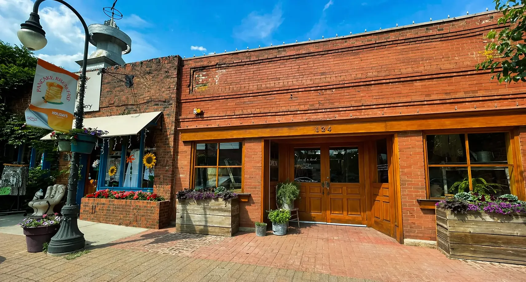 Brick storefront with large wooden door and windows, decorated with potted plants and flowers outside, a lamppost with hanging sign and decorations, and a blue sky with some clouds.