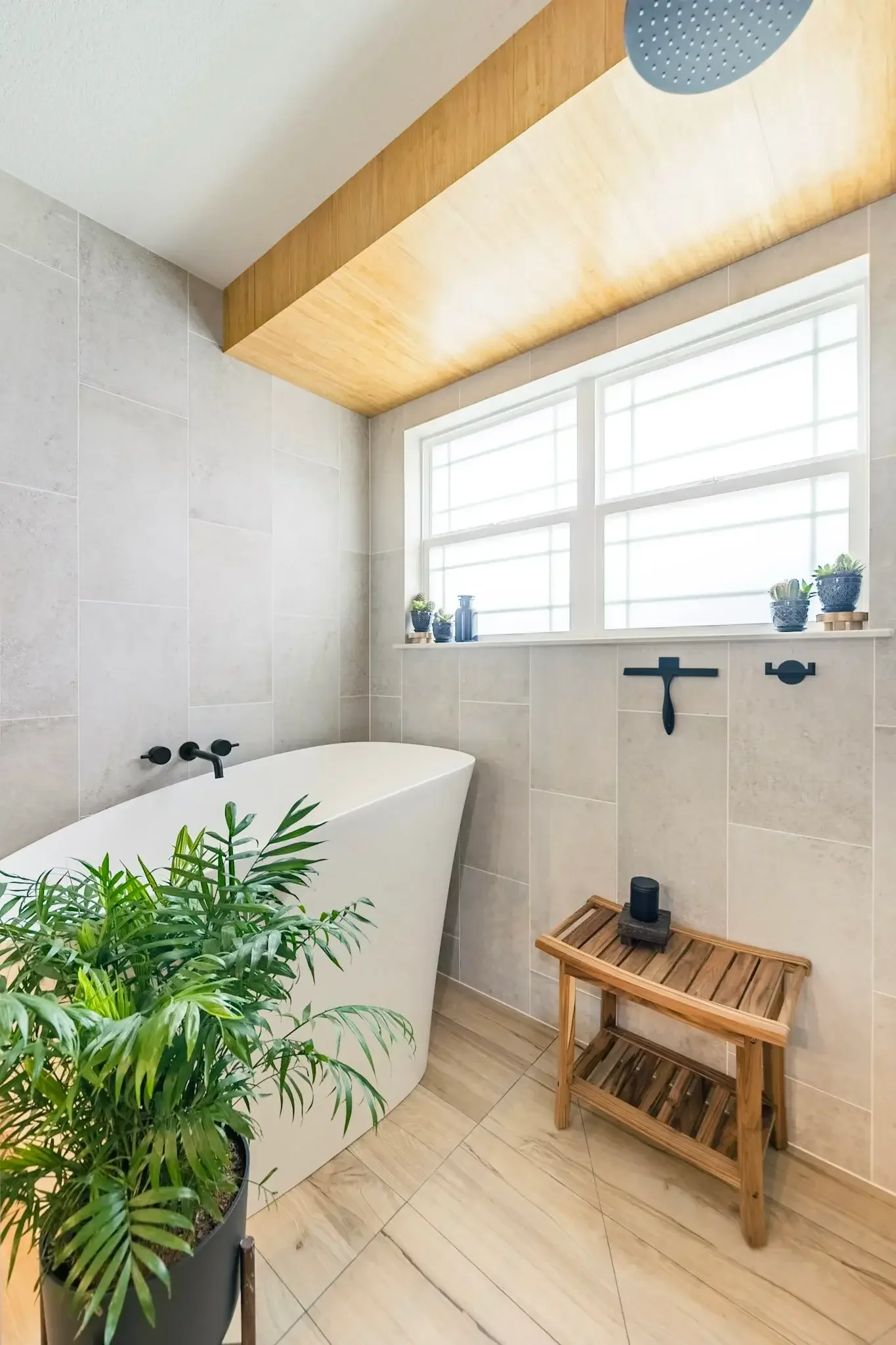 A modern bathroom with a white bathtub, large beige tiled walls, a large window with white framing, plants on the windowsill, a wooden ceiling accent, a plant in a black pot, and a wooden towel rack with a black soap dispenser on top.