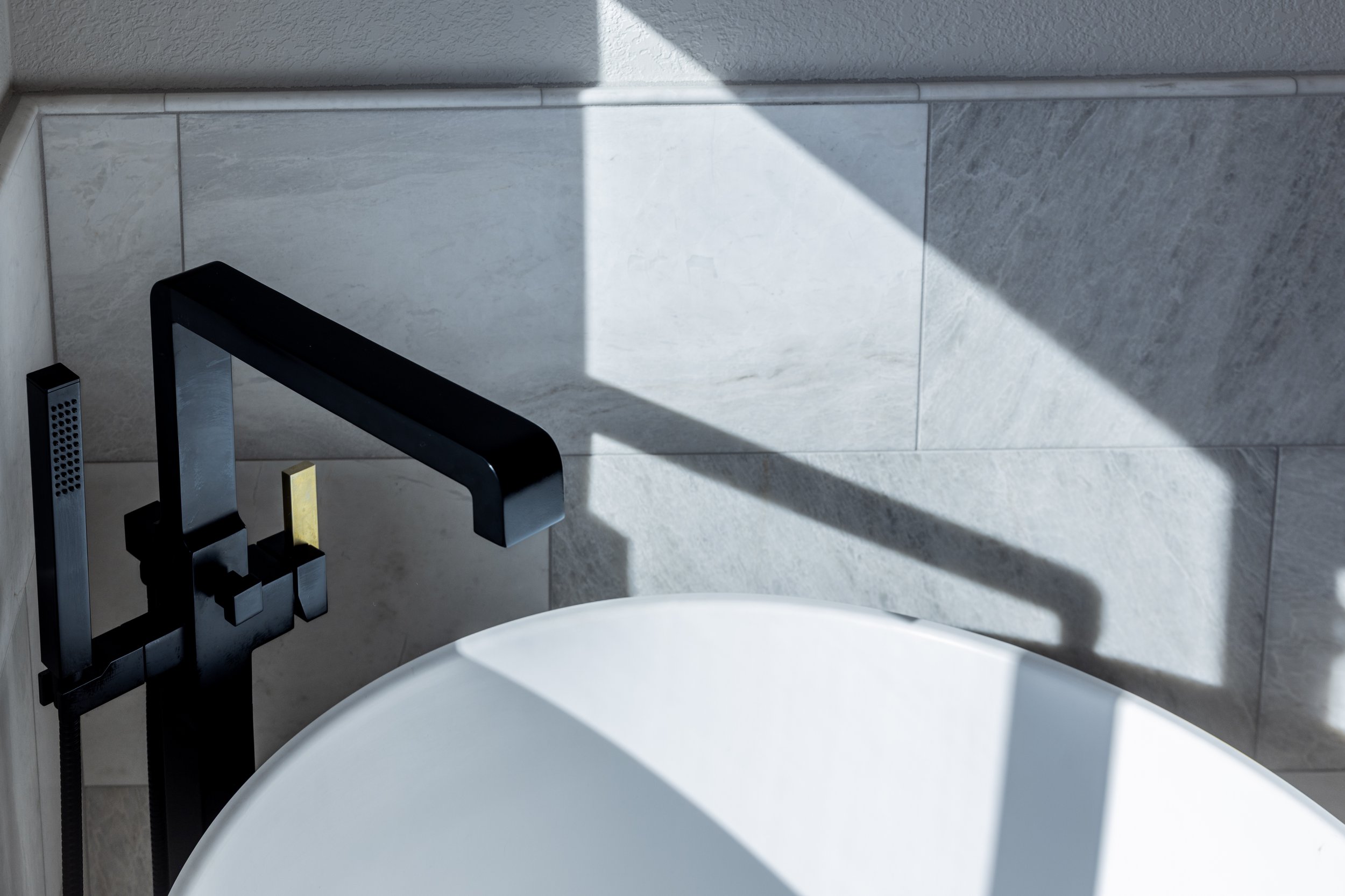 Close-up of a modern black faucet beside a white bathtub, with a tiled wall in the background and shadows cast by sunlight.