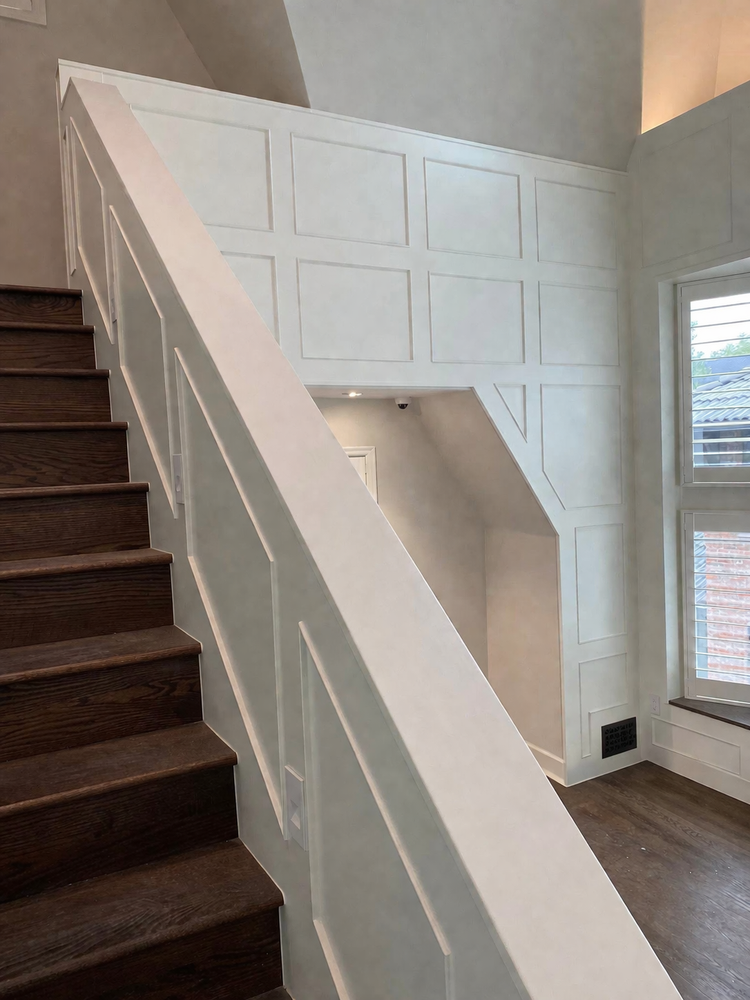 Interior view of staircase with dark wooden steps and white railing, adjacent to a wall with decorative paneling and windows.
