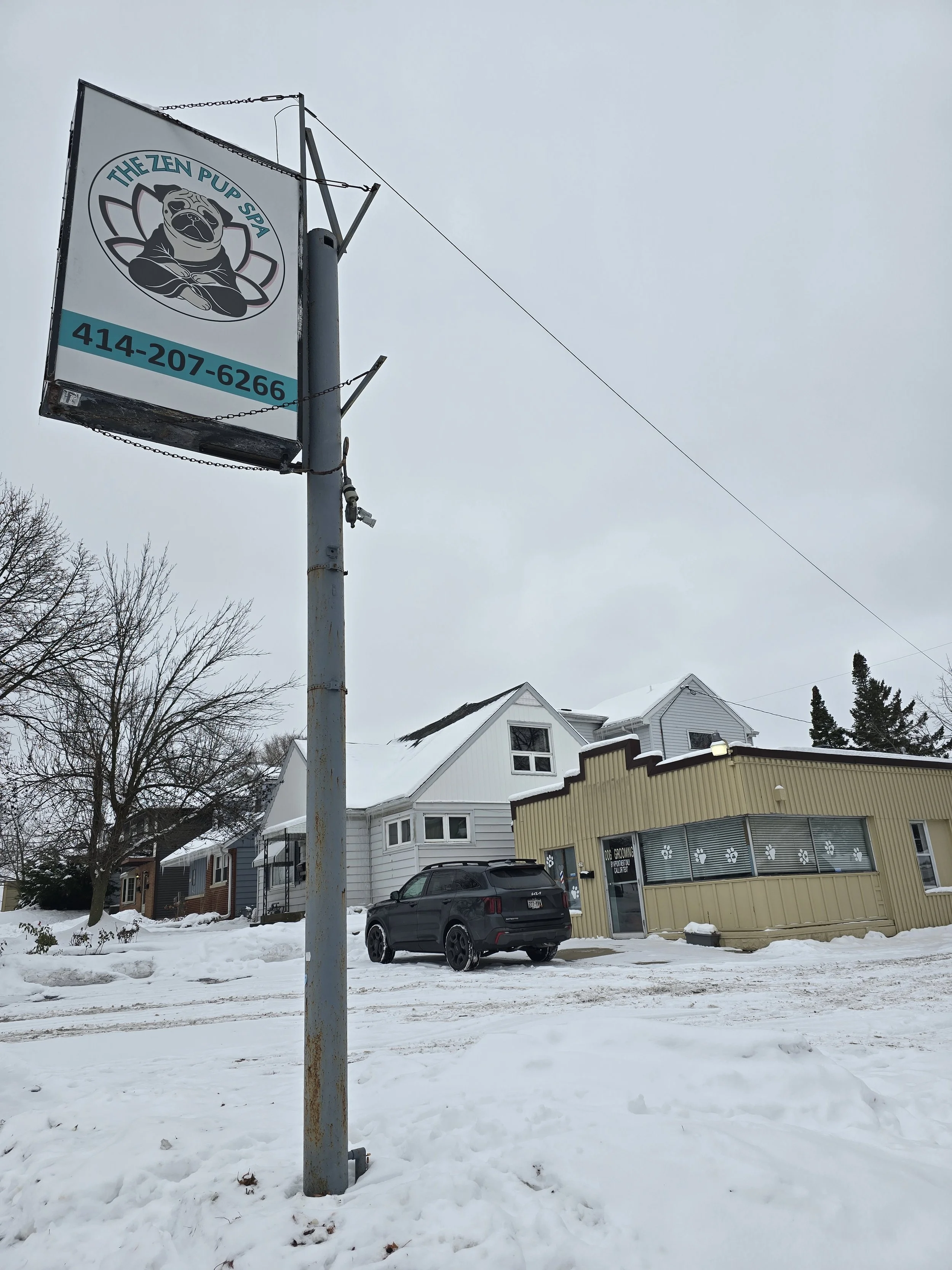 Sign for The Zen Pup Spa with a cartoon dog logo and phone number, set against a snowy street with houses and a car in the background.