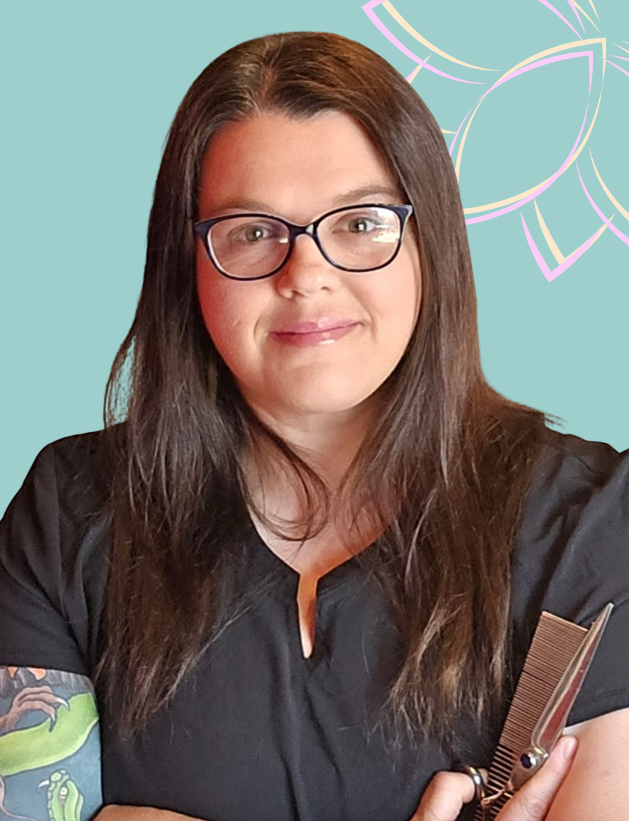 A woman with long brown hair, glasses, and a black shirt holding a pair of hair scissors with a comb in her hand. She is smiling in front of a colorful abstract background.