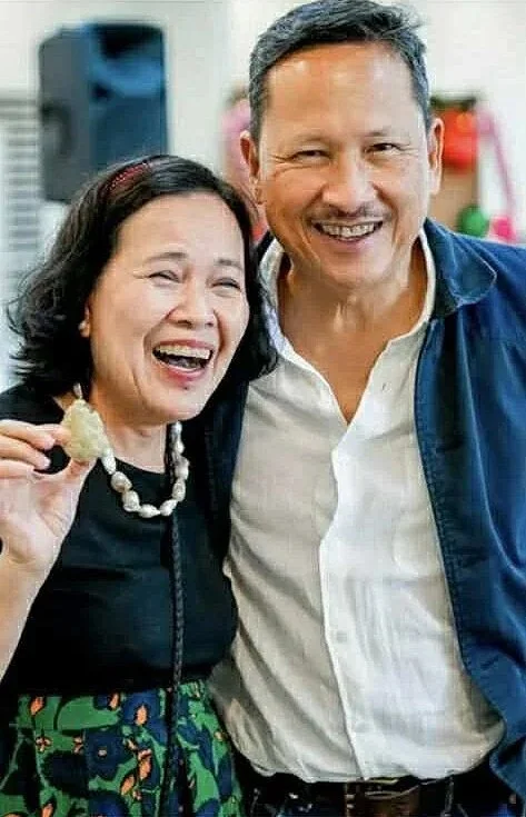 A smiling woman and man standing close together, the woman holding a piece of food, in a festive or social setting.