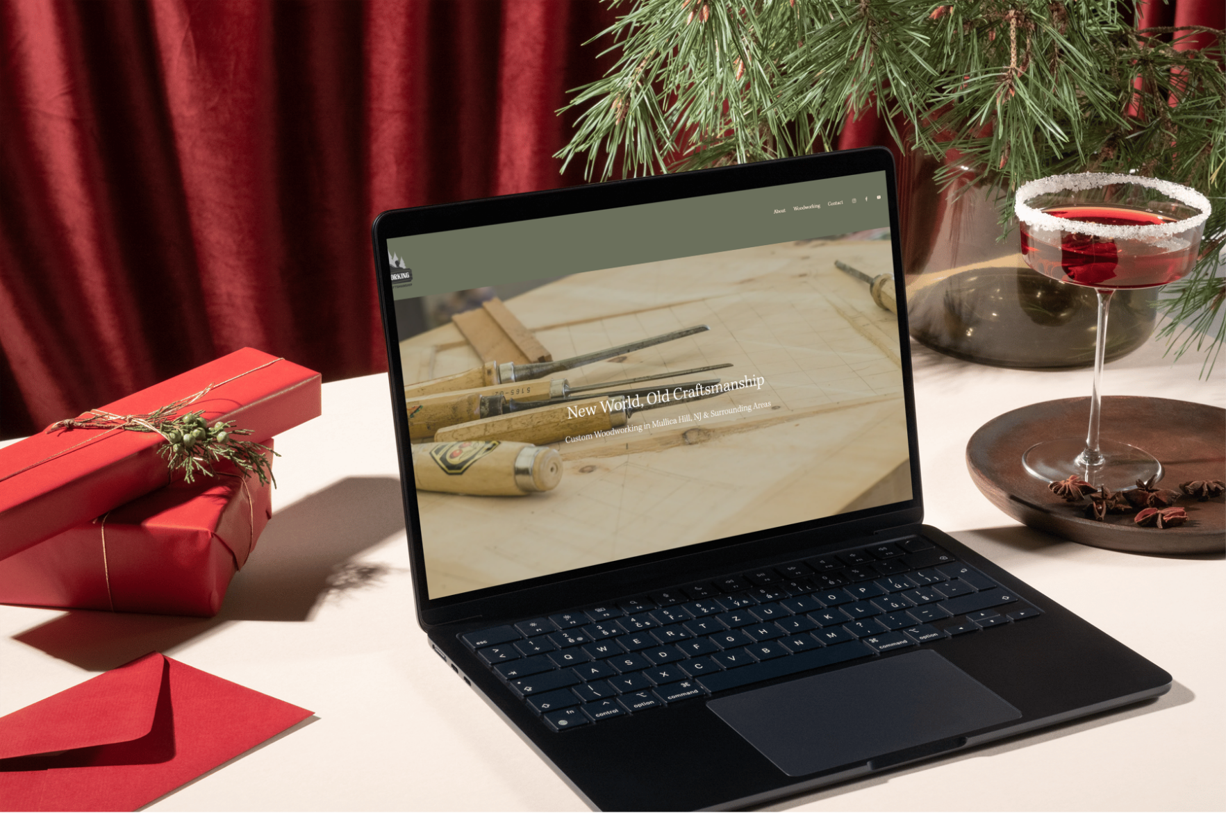 Laptop on a white table displaying a woodworking website, surrounded by Christmas decorations including a red gift box with a sprig of pine, a red envelope, a glass of red wine with a salted rim on a dark plate, a black vase with pine branches, and a red curtain in the background.