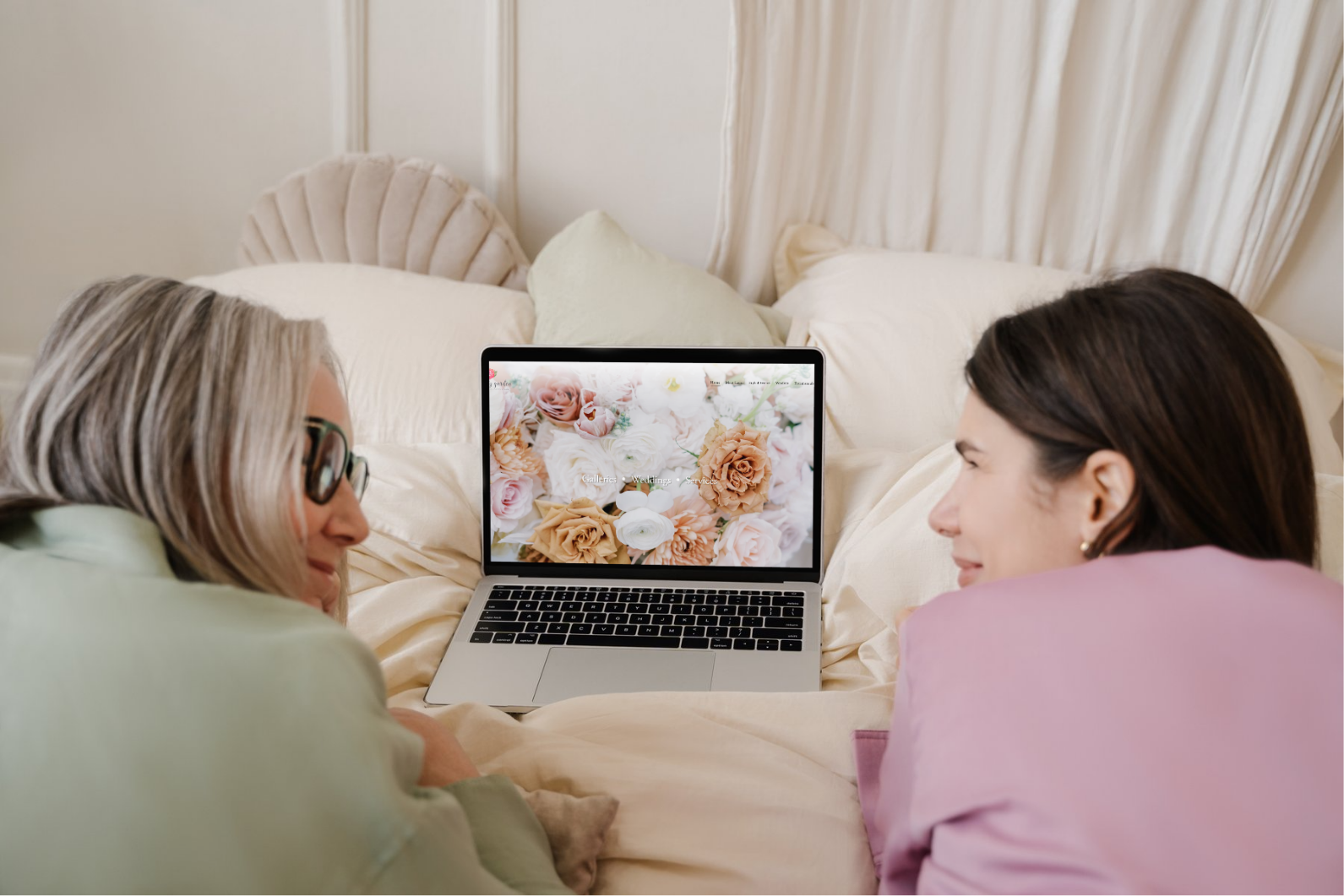 Two women lying on a bed, looking at a laptop with a floral website displayed.