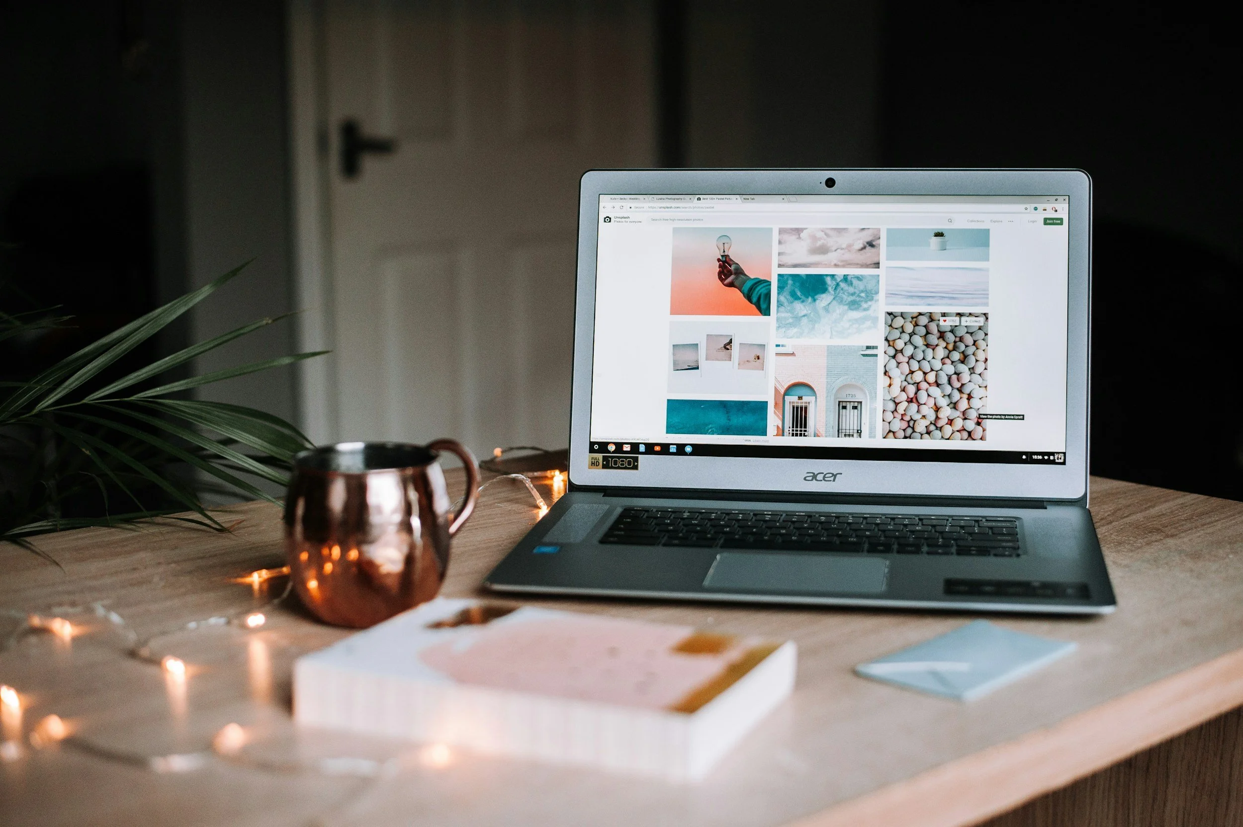 A laptop on a wooden desk displaying a grid of digital photos, next to a copper mug, a book, and a smartphone. Decorative string lights and a green plant are also on the desk.