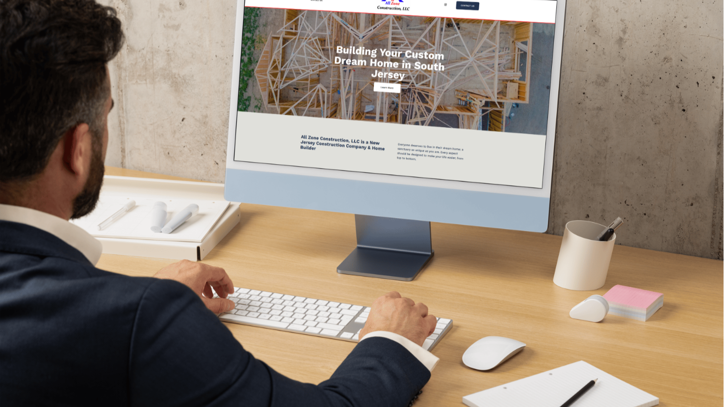A man in a suit using a desktop computer to view a construction company's website in an office with a concrete wall. The website shows a wooden house frame and text about building a custom home in South Jersey.