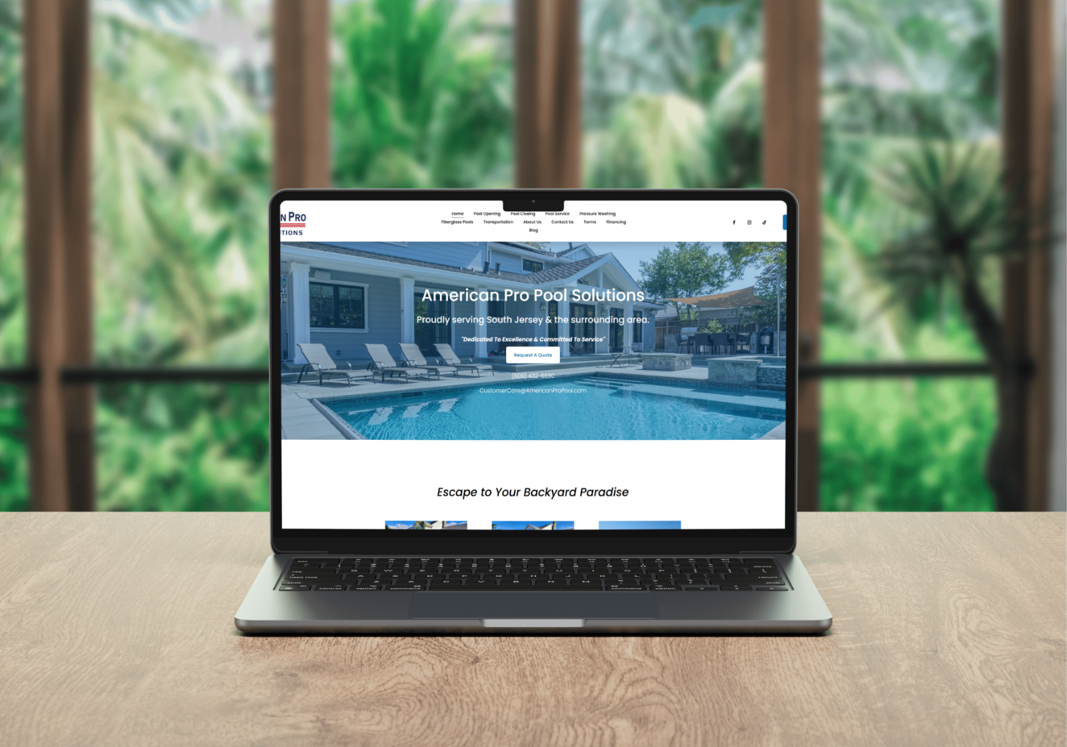 Open laptop on a wooden table displaying the homepage of American Pro Pool Solutions, featuring a backyard pool with lounge chairs and patio furniture, with a blurred view of trees and outdoor scenery in the background.