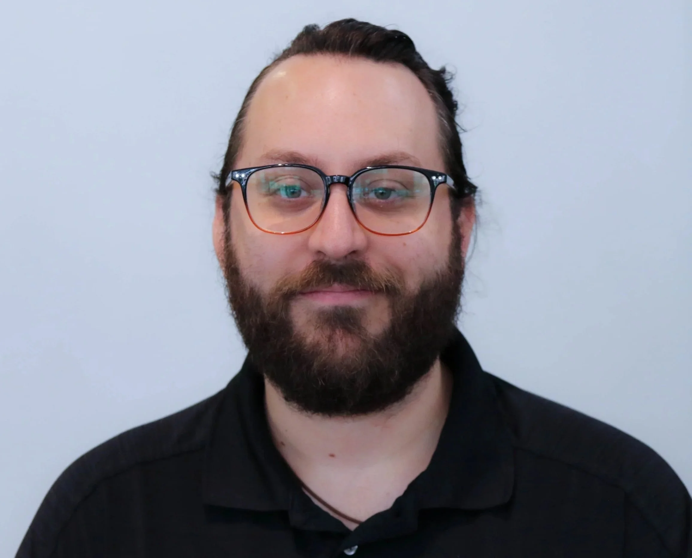 Close-up of a man with glasses and a beard, wearing a black shirt, against a plain white background.