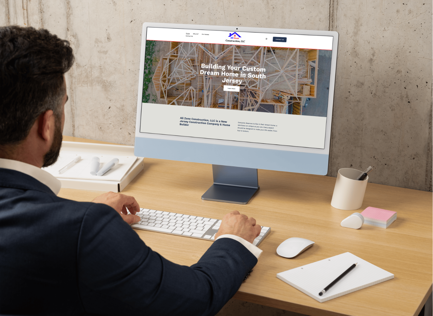 A man in a suit working at a wooden desk with a computer displaying a construction company's website, surrounded by office supplies.