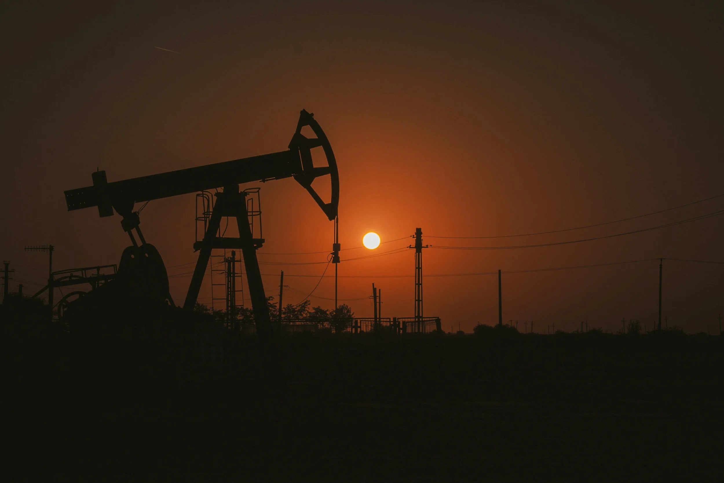 Silhouette of an oil pumpjack against a sunset sky with a setting sun and power lines.
