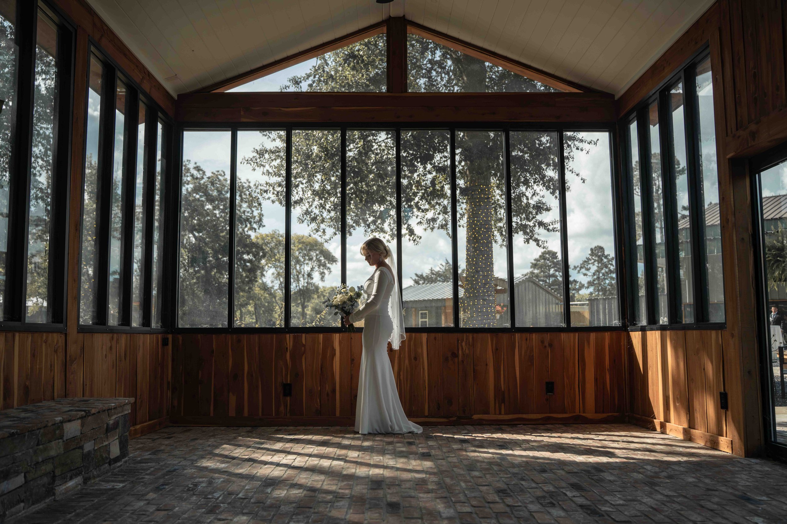 Bride holding a white floral bouquet inside the glass-walled Bloom House at Mimosa Gardens in Dayton, Texas, with natural light and garden views.