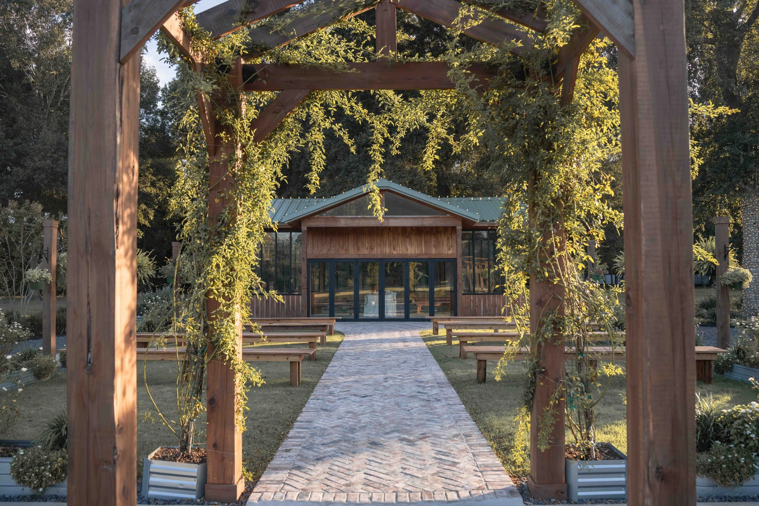 Wooden wedding ceremony arbor surrounded by greenery, with a brick aisle, guest benches, and the Bloom House at Mimosa Gardens wedding venue in Dayton, Texas.