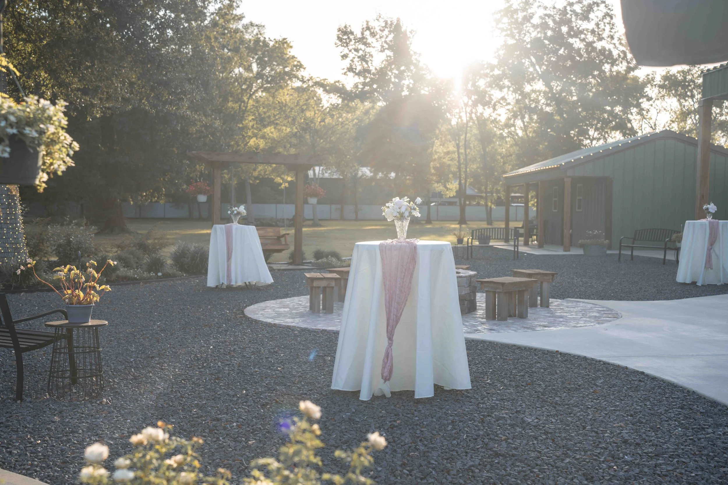 Outdoor wedding cocktail hour at Mimosa Gardens in Dayton, Texas, featuring floral arrangements on cocktail tables beneath the trees in warm evening sunlight.