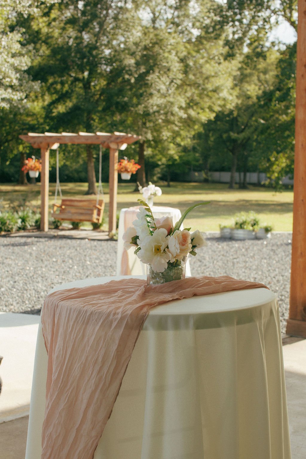 Outdoor wedding reception table decorated with a blush pink runner and white floral centerpiece, overlooking the garden and wooden swing at Mimosa Gardens in Dayton, Texas.