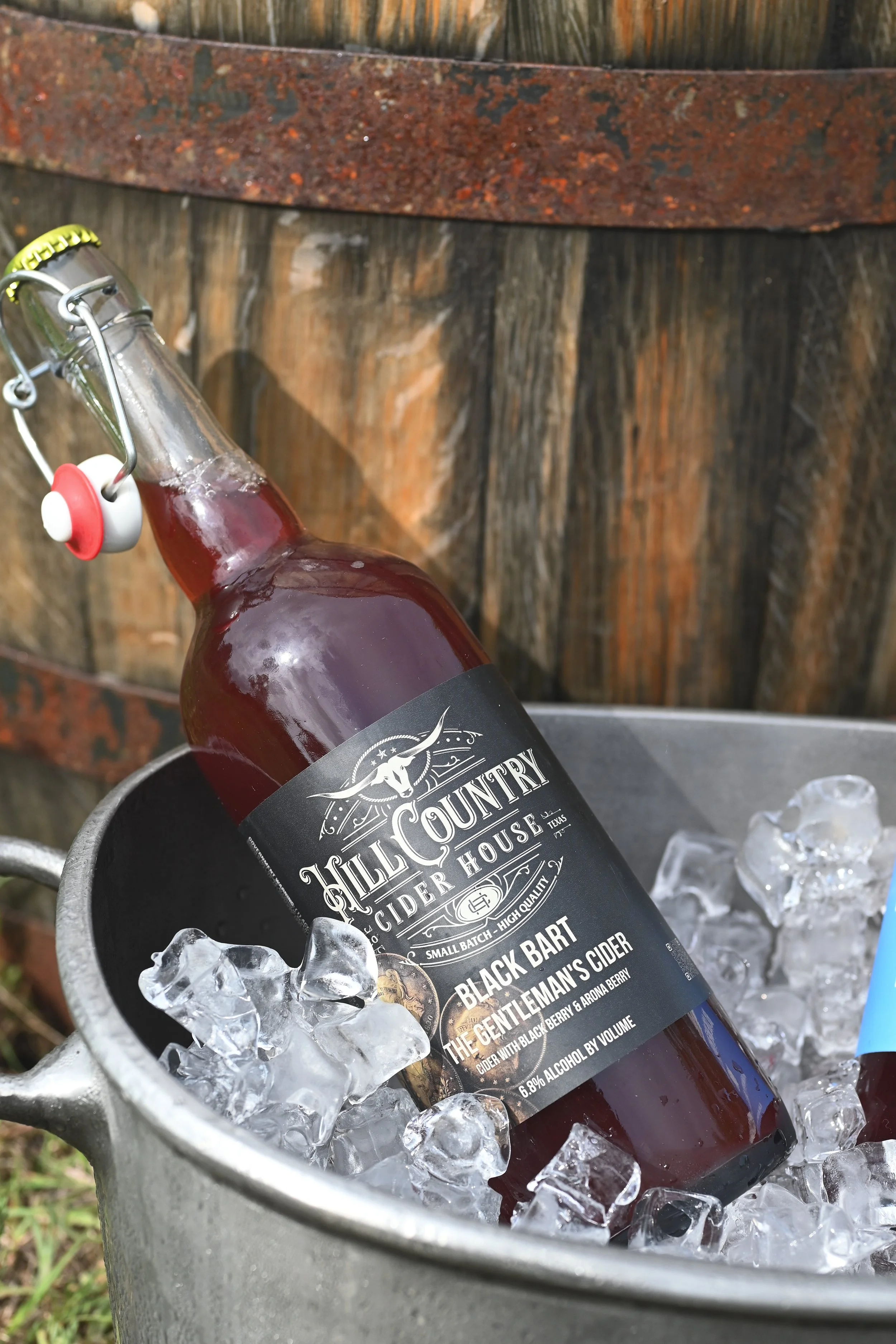 A bottle of Hill Country cider, Black Bart, sitting on ice in a metal bucket outdoors, with a wooden fence in the background.