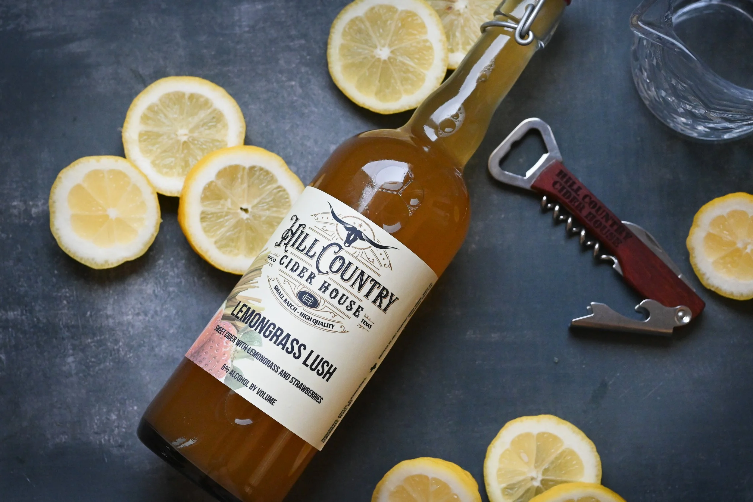 A bottle of Hill Country cider with a lemon flavor, surrounded by lemon slices and a glass water pitcher on a dark surface, with a bottle opener nearby.