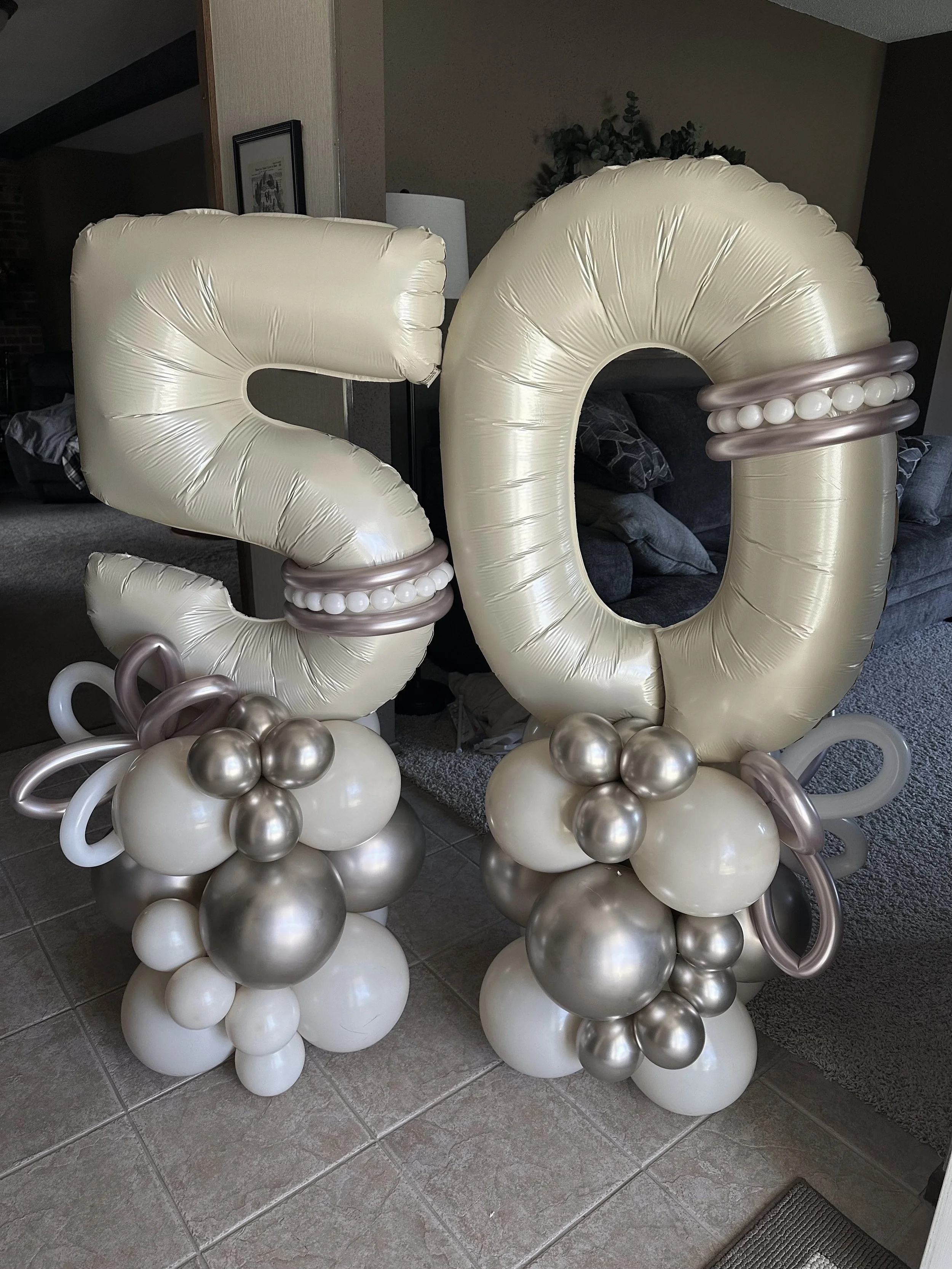 Large number '20' made of cream, silver, and metallic balloons, decorated with a pearl-like balloon ring, placed indoors on a tiled floor.