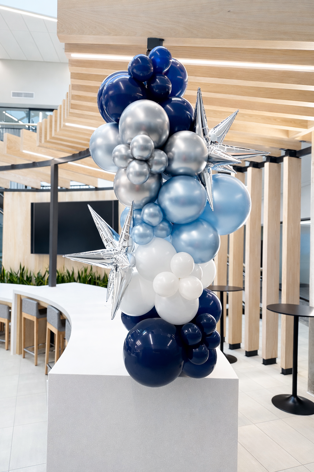 Balloon arrangement in shades of blue, silver, and white with star-shaped metallic balloons, set in a modern indoor space with wood paneling and black tables.
