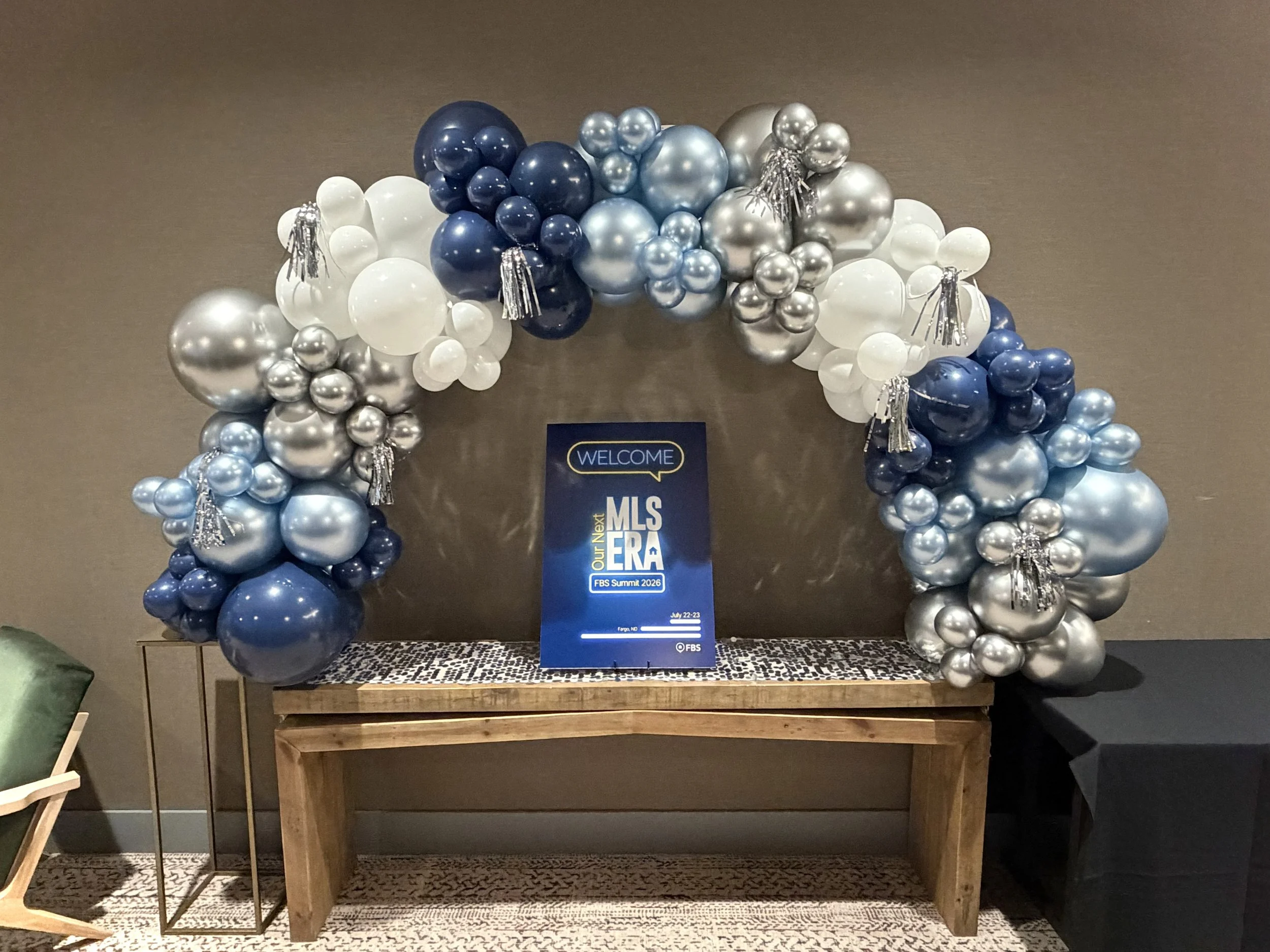 Balloon arch in blue, silver, and white with metallic and pearl finishes. Some balloons have tassel and fringe decorations. A sign with 'Welcome' and 'MLS Era' is underneath the arch on a wooden table.
