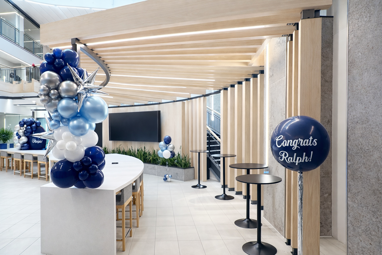 Decorations for a celebration, including blue, white, and silver balloons, a 'Congrats Ralph!' balloon, and a long white table with chairs in a modern indoor space.