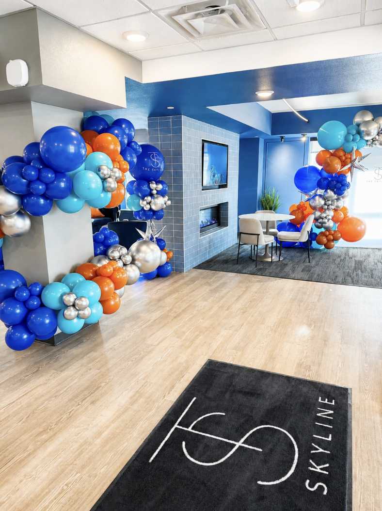 Decorated interior with colorful balloons in blue, orange, teal, and silver, arranged along walls in a modern space. The room features a fireplace, a television, a small table with chairs, and a logo on a rug that reads 'SKYLINE.'